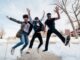 photo of three men jumping on ground near bare trees during daytime