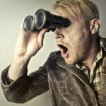 A surprised man in a leather jacket peers through binoculars with a shocked expression.