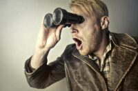 A surprised man in a leather jacket peers through binoculars with a shocked expression.