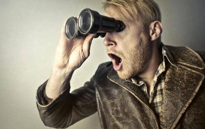 A surprised man in a leather jacket peers through binoculars with a shocked expression.