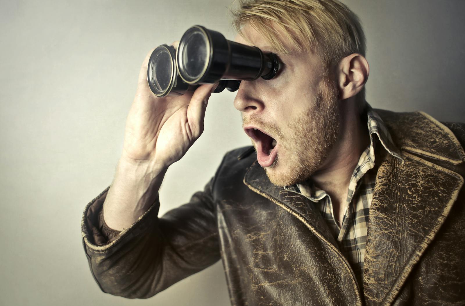 A surprised man in a leather jacket peers through binoculars with a shocked expression.