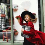 Fashionable woman in a red coat walks by a stylish boutique window, carrying a magazine.