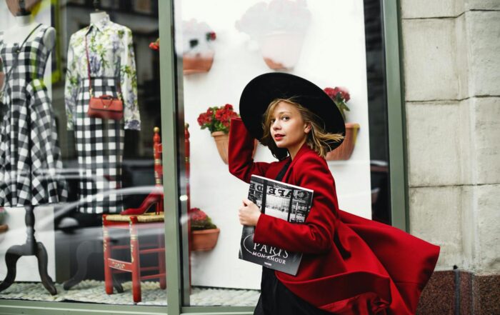 Fashionable woman in a red coat walks by a stylish boutique window, carrying a magazine.