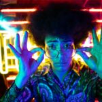 A young adult in glasses poses with neon lights, showcasing retro fashion and vibrant nightlife vibes.