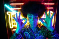 A young adult in glasses poses with neon lights, showcasing retro fashion and vibrant nightlife vibes.