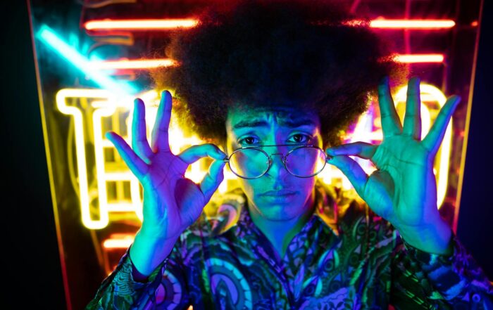 A young adult in glasses poses with neon lights, showcasing retro fashion and vibrant nightlife vibes.