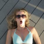 Blonde woman in sunglasses lying outdoors on a wooden deck, looking surprised.
