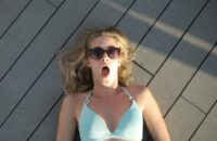 Blonde woman in sunglasses lying outdoors on a wooden deck, looking surprised.