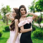 Two women in formal dresses pose outdoors in a lush garden setting, radiating elegance.