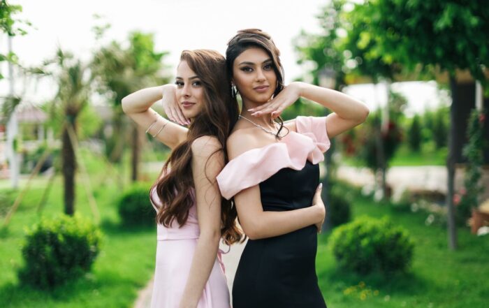 Two women in formal dresses pose outdoors in a lush garden setting, radiating elegance.